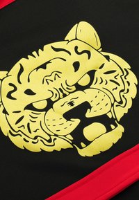 Schwarzer Stoff mit einem auffälligen Design eines gelben Tigergesichts, schwarzen Details und einem roten Akzent am unteren Rand.