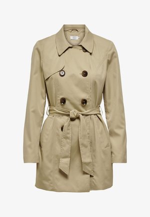 Mujer que lleva un abrigo trench beige de doble botonadura con cinturón, suéter azul, vaqueros azules de pierna recta y botines negros, de pie con una mano en el bolsillo.