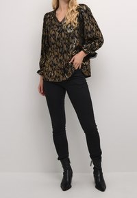 Blouse noire et dorée à motifs avec un col en V et des manches ballons, associée à un jean skinny noir et des bottines. Tissu texturé, accents en dentelle.