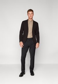 Blazer nero sopra una camicia beige con bottoni, abbinato a pantaloni grigi e scarpe nere. Tessuto liscio, design strutturato, vestibilità versatile.