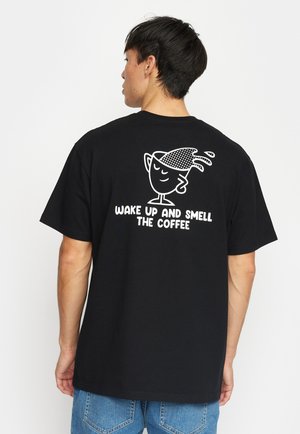 Zwarte katoenen t-shirt met een speelse graphic van een koffiekop en de tekst "WORD WEKKER EN RUIK DE KOFFIE" op de achterkant. Korte mouwen, ontspannen pasvorm.