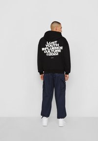 Svart hettegenser med hvit tekst på baksiden som leser "LOST YOUTH INFLUENCE CULTURE ©2022." Kombinert med marineblå cargo-bukser og hvite sneakers.