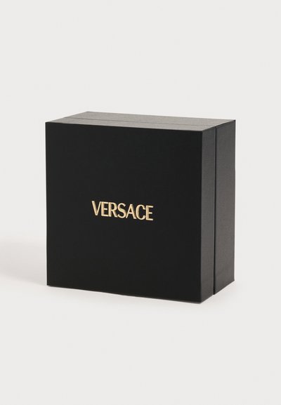 Μαύρο ορθογώνιο κουτί δώρου Versace με χρυσή ανάγλυφη επωνυμία στη μπροστινή πλευρά, εμφανιζόμενο σε απλό φωτεινό φόντο.