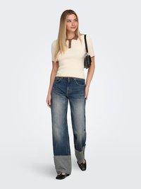 Žena s dugom plavom kosom nosi bež kratku majicu, dvobojne široke traperice s manžetnama, crne cipele i nosi crnu torbu preko ramena.