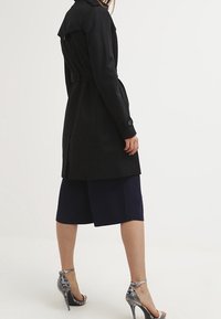 Abrigo de trench negro con cintura ceñida, diseño ajustado, de pie en paralelo al espectador, combinado con pantalones cortos azul marino y tacones altos plateados.