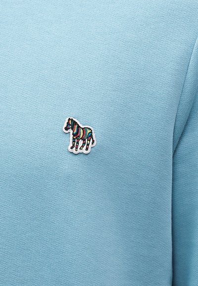 Ανοιχτό μπλε ύφασμα με ένα μικρό κεντημένο πατς ζέβρας που διαθέτει πολύχρωμες ρίγες στην περιοχή του στήθους.