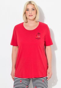 T-shirt rouge avec un col rond, ornée d'une broderie de cerises noires et de texte. Ensemble porté avec un pantalon rayé noir et blanc avec un motif de cerises.