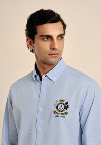 Camisa de botones a rayas de color azul claro, hecha de una tela de textura suave; presenta un logotipo náutico bordado en el pecho izquierdo.