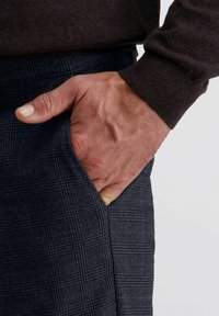 Volwassen hand gedeeltelijk in de zijzak van donkergrijs geruite broek, met een donkerbruine lange mouwen trui aan.
