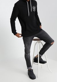 Black hooded sweatshirt with white drawstrings, front pocket, and graphic design. Paired with gray distressed jeans and black sneakers.