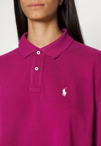 Magenta polo tričko s klasickým límcem, dvěma knoflíky a bílým logem na prsou. Materiál má strukturovaný pletený povrch.