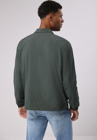 Tmavě zelená lehká bunda s límcem, elastickými manžetami a volným střihem, nošená přes světle modré džíny. Hladká textura.