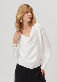 Biała bluzka z luźnym dekoltem, długimi rękawami i miękką fakturą, zestawiona z ciemną spódnicą i złotymi bransoletkami.