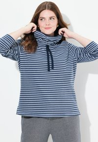Pullover a righe blu e bianche con collo a camicia, maniche 3/4, dettaglio con coulisse e vestibilità comoda. Indossato con pantaloni grigi a fantasia.