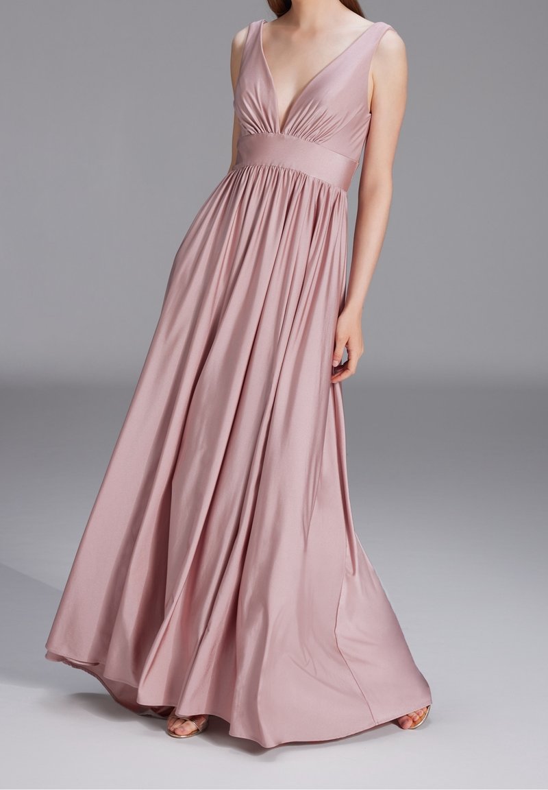 Femme portant une robe longue sans manches rose poudré avec un profond décolleté en V et une taille froncée, debout devant un fond gris uni.