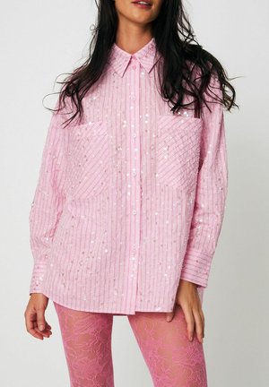 Žena v voľnej ružovej košeli na gombíky s trblietkami a ružových kvetinových čipkovaných pančuchách stojaca pred jednoduchým pozadím.