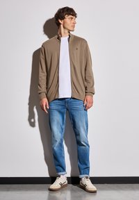 Hnědá zipová bunda přes bílý tričko, spárovaná se světle modrými džíny a krémovými teniskami. Bunda má stojáček a žebrované manžety.