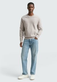 Junger Mann steht posierend in einem beigen Pullover, hellblauen Jeans und weißen Sneakers, mit einer Hand in der Tasche vor einem schlichten Hintergrund.