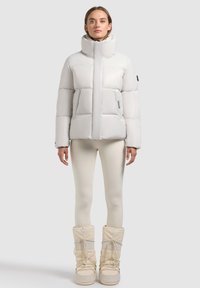 Lys gray puffer jakke med høj krave, firkantet design og quiltet tekstur; parret med cremefarvede leggings og hvide støvler.