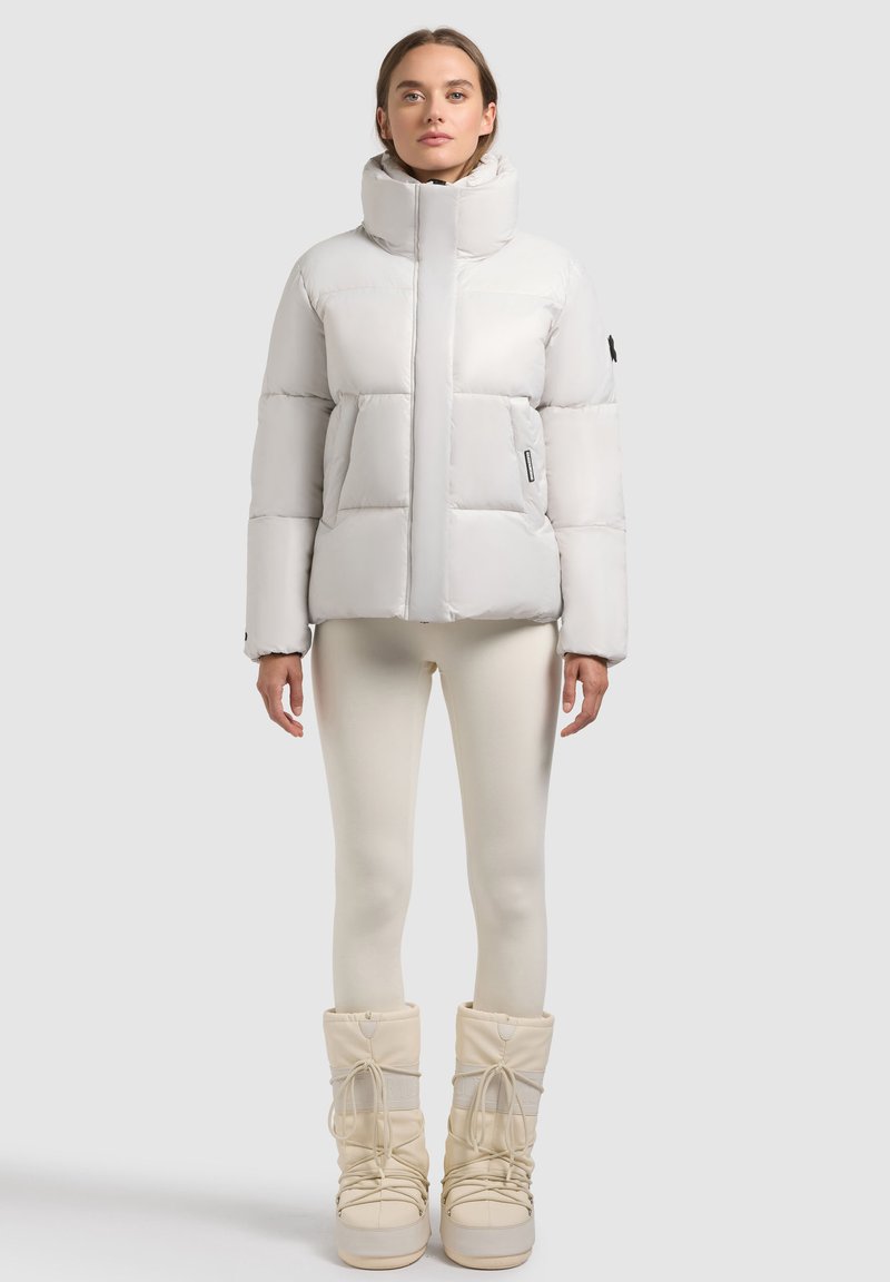 Lys gray puffer jakke med høj krave, firkantet design og quiltet tekstur; parret med cremefarvede leggings og hvide støvler.