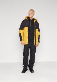 Chaqueta acolchada en negro y amarillo con capucha, que cuenta con un cierre frontal, bolsillos en el pecho y laterales, combinada con pantalones cargo negros y botas.
