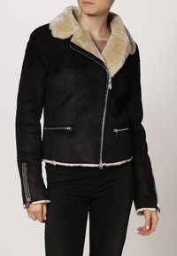 Svart mockajacka med en beige fuskpälskrage, silverfärgade dragkedjor och dragkedjefickor. Croppad design med pälsdetalj vid nederkanten.