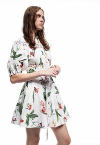 Witte bloemenjurk met pofmouwen en een gecinchte taille, voorzien van kleurrijke bloemen en groene bladeren in verschillende patronen, knielange snit.
