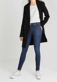 Cappotto di lana nero con design doppiopetto, t-shirt grigia, jeans skinny blu e sneakers bianche. Sfondo neutro con illuminazione soffusa.