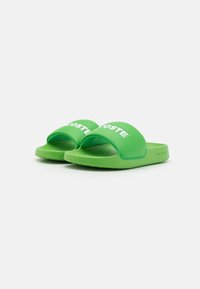 Sandales à滑绿色 en caoutchouc avec une large lanière, présentant le logo "LACOSTE" en blanc. Semelle intérieure texturée pour adhérence et confort. Semelle plate.
