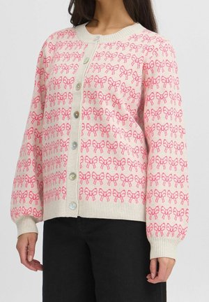 Woman wearing a cream cardigan with pink bow patterns, buttoned down the front, paired with black pants.