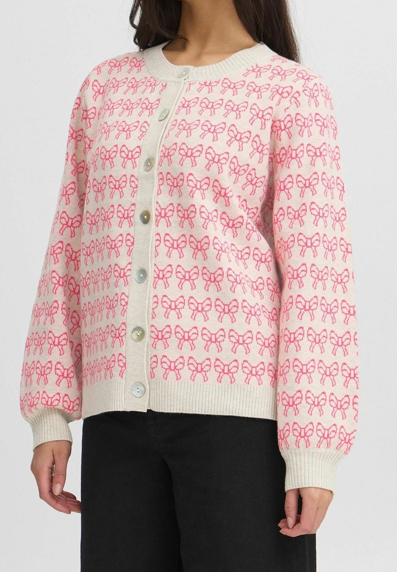 Femme portant un cardigan crème avec des motifs de nœuds roses, boutonné sur le devant, associé à un pantalon noir.