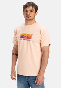 Světle broskvové tričko vyrobené z bavlny, se barevnou grafikou loga Quiksilver umístěnou na hrudi, s klasickým kulatým výstřihem.