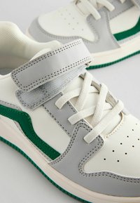 Hvide og grå sneakers med grønne accenter, der har en tekstureret rem, perforeret tå og syntetiske materialer. Snøret med flade hvide snørebånd.
