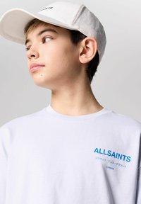 Balta medvilninė marškinė su mėlynu "ALLSAINTS" spaudiniu kairėje krūtinės pusėje, derinama su šviesių spalvų kepure, turinčia kreiptą vizorą.