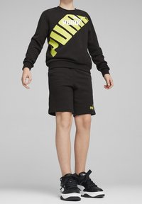 Felpa nera con grande logo Puma giallo, abbinata a pantaloni corti neri coordinati. Indossata con sneakers nere con accenti bianchi.