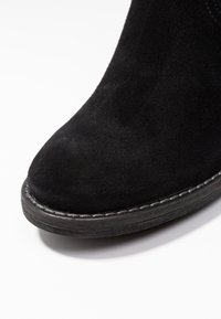 Bottine en daim noir avec un bout arrondi, une finition texturée et une semelle plate noire ornée de coutures blanches sur le bord.