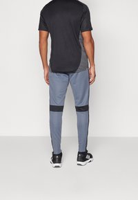 Grijze sportbroek met zwarte zijkanten, gecombineerd met een zwart shirt met korte mouwen met grijze accenten. Draagt zwarte sportschoenen.