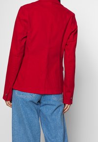 Červený bavlněný blazer s přiléhavým střihem, dlouhými rukávy a zapínáním na jeden knoflík. Spárovaný se světle modrými denimovými kalhotami.