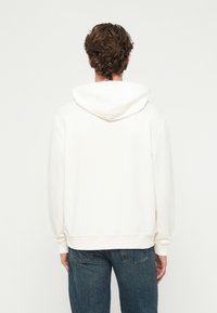 Hanorac alb cu croială lejeră, glugă cu șnur și manșete striate. Asortat cu blugi bleumarin, cu o textură ușor uzată.