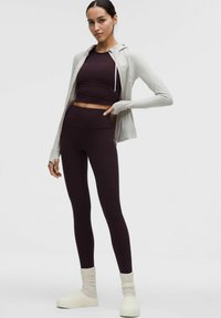 Atletska oprema, ki vključuje prilegajoč temno vijoličen top, ujemajoče se hlače z visokim pasom, svetlo sivo zadrgo s kapuco in bele slip-on čevlje z teksturiranimi nogavicami.