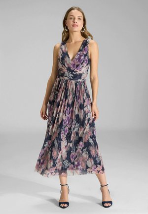 Femme portant une robe fleurie sans manches, col en V, mi-longue, dans des tons violet, rose et bleu, assortie à des talons à bride cheville bleu marine, debout sur un fond gris.