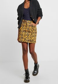 Cardigan negro de punto sobre una blusa azul marino, combinado con una falda amarilla de estampado de serpiente. Modelo con botas negras de cordones.