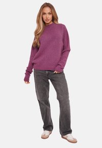 Purple knit sweater with long sleeves, relaxed fit. Paired with gray denim jeans and white sneakers, standing with one hand on hip.