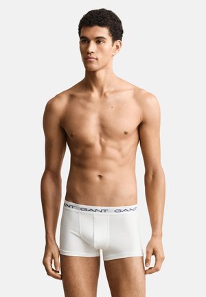 Junger Mann mit kurzen lockigen Haaren, der weiße GANT Boxershorts trägt, steht vor einem einfarbigen hellen Hintergrund und blickt leicht nach rechts.