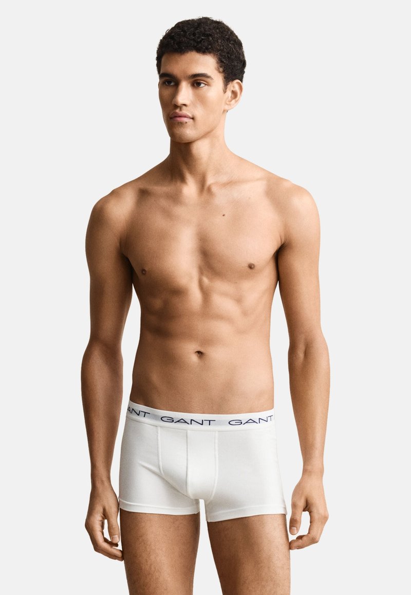 Jeune homme aux cheveux courts et bouclés, portant un boxer blanc GANT, debout devant un fond clair uni, regardant légèrement vers la droite.