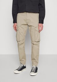 Man in beige cargo joggerbroek met elastische manchetten, zwart langemouwen shirt en zwarte lage sneakers met witte veters.