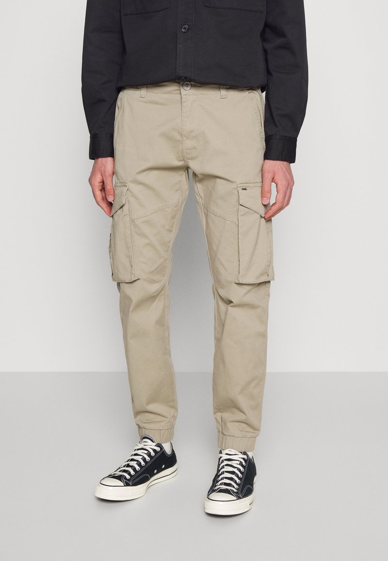 Man in beige cargo joggerbroek met elastische manchetten, zwart langemouwen shirt en zwarte lage sneakers met witte veters.