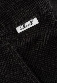 Black corduroy fabric with a textured pattern. A white label showing the brand name "Reell" is partially visible along the edge.