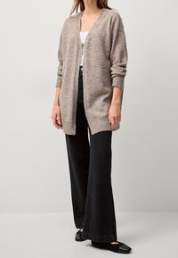 Jasnobrązowy kardigan, otwarty z przodu, z prążkowanymi mankietami i dołem. Połączony z czarnymi spodniami w szerokim kroju i płaskimi butami, na neutralnym tle.