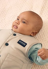 Veste matelassée beige en tissu léger, avec une étiquette bleu foncé et des boutons noirs. En dessous, une chemise à manches longues bleu clair.